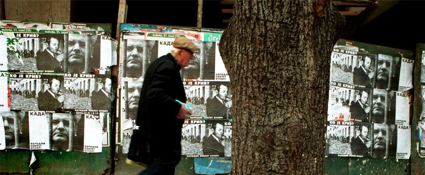 An image of an old man walking past a wall plastered over with news pamphlets.