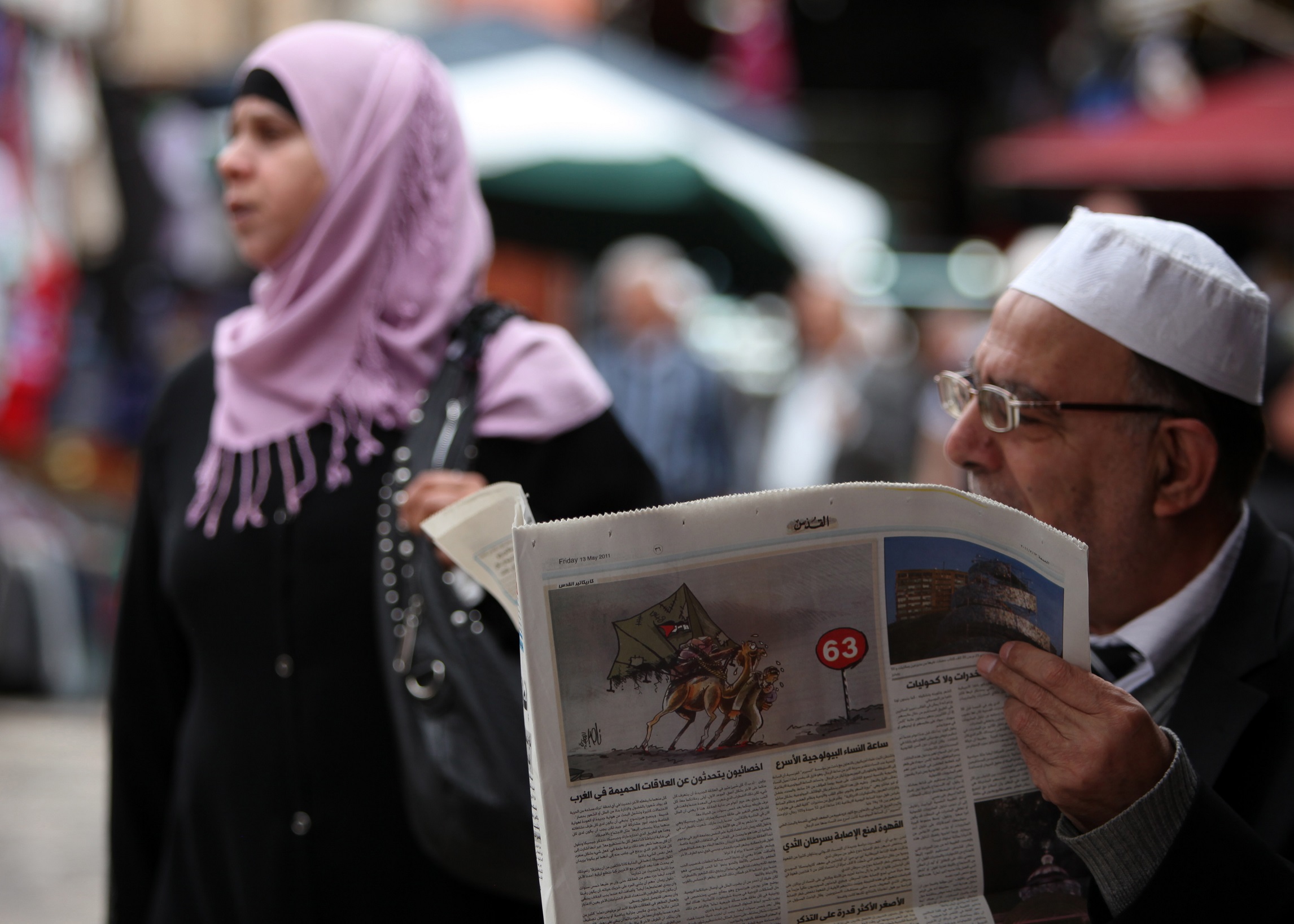 A picture of a man in glasses reading the paper while a woman in a pink hijab walks behind him, not looking at the camera. 