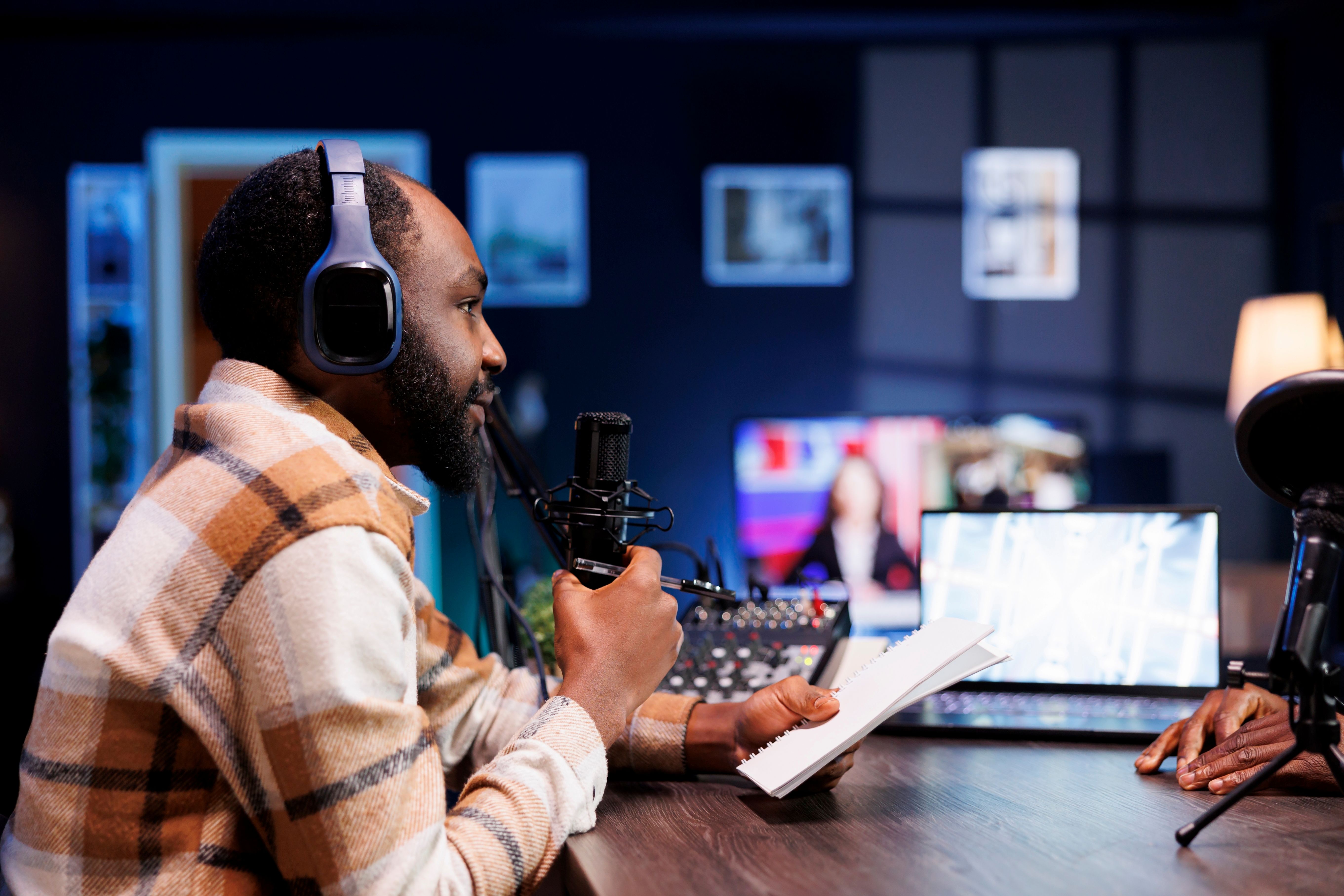 African male content creator is hosting an online radio show and talking into a microphone. It is a podcast episode in which the host, a black guy, does an online interview at his home studio.