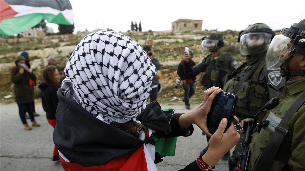 ناشطة فلسطينية تلتقط صورة للقوات الإسرائيلية خلال احتجاج أسبوعي ضد المستوطنات اليهودية، في قرية النبي صالح بالضفة الغربية. تصوير: محمد توركمان – رويترز.