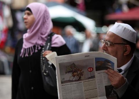 A picture of a man in glasses reading the paper while a woman in a pink hijab walks behind him, not looking at the camera. 