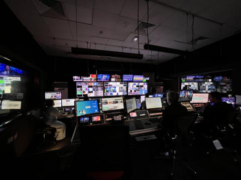 Al Jazeera English News Control-room. A dimly lit newsroom control room with multiple operators seated at large desks, surrounded by numerous monitors and screens displaying live news feeds, control panels, and broadcasting software. The central wall is covered with a grid of television screens showing various live broadcasts and graphics, creating a high-tech, high-pressure environment.