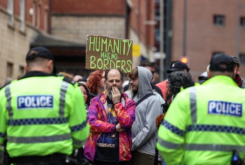 A man standing in the center of a protesting crowd, thinking, with a placard behind him that reads "Humanity has no borders"