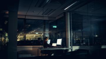 A man in a dark office room is working on a computer with two screens.